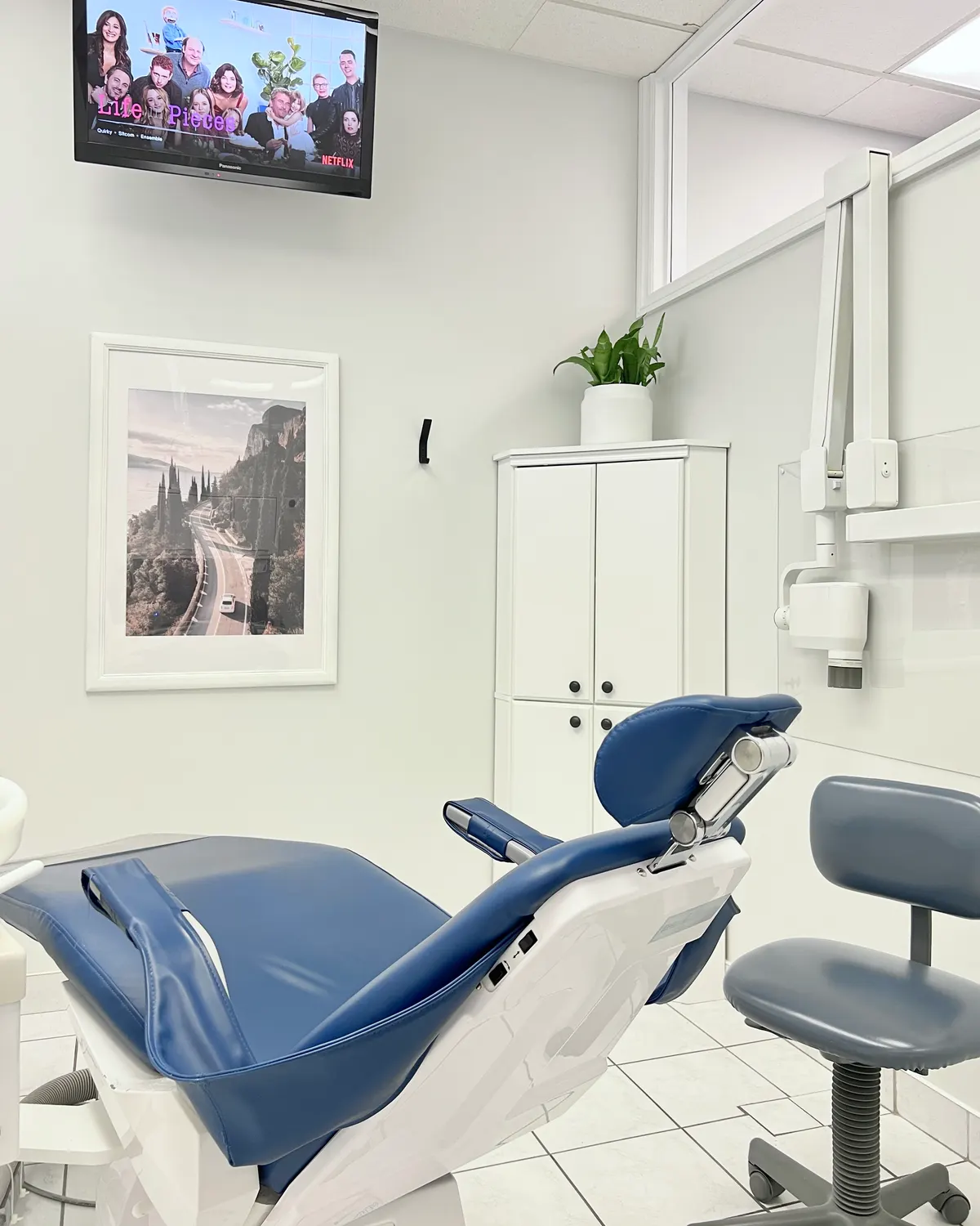 Headon treatment room with a navy dental chair, rolling stool, a framed print and a wall-mounted TV