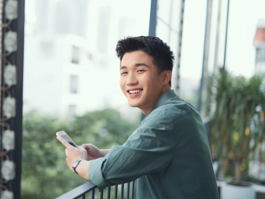 A smiling young man holding his phone on a balcony